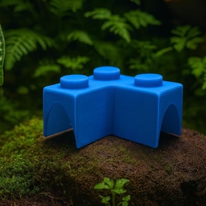 May include: A vibrant blue building block, featuring three studs on top and an arched design. The object is set against a backdrop of green foliage and moss. The block's design is reminiscent of a classic toy.