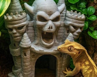 Op de afbeelding: Een grijs, kasteel-vormig reptielenverblijf met een dreigend schedelontwerp, compleet met torentjes en een grote open mondingang. Een geel-bruine gekko is gepositioneerd in de buurt van het kasteel. De achtergrond heeft groen gebladerte.