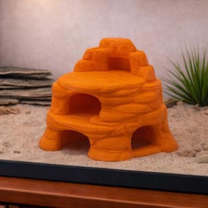 May include: An orange, layered rock formation with multiple openings, designed as a habitat for small pets. The textured surface mimics natural rock, providing a visually appealing and functional shelter. The structure is placed in a terrarium setting.