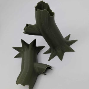 May include: Two olive green, stylized tree trunk sculptures with star-shaped bases. The trunks have a rough, textured appearance with jagged edges and a hollowed-out top, suggesting a broken or weathered aesthetic. The sculptures are likely decorative.