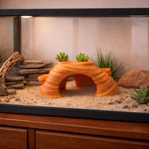 May include: An orange resin reptile hide with an arched opening and two small succulent plants on top. The hide is in a terrarium with sand, rocks, and other decorative elements. The terrarium is set on a wooden cabinet.