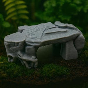 Op de afbeelding: Een grijze, miniatuur stenen structuur die lijkt op een kleine tafel of altaar. Het tafelblad heeft gedetailleerde oppervlaktestructuren en de structuur wordt ondersteund door twee pilaren. Het object is waarschijnlijk een decoratief stuk of een onderdeel voor een miniatuurscène.