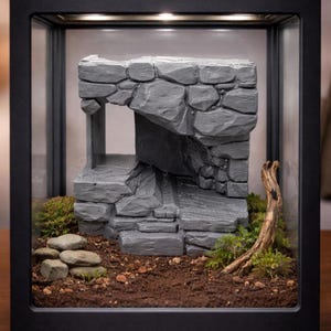 May include: A miniature grey stone structure with an archway and steps, displayed in a black framed glass case. The diorama includes soil, small rocks, moss, and a piece of driftwood, creating a naturalistic scene.