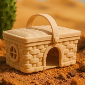 Puede incluir: Una estructura beige en forma de cesta con asa, ventana redonda y puerta arqueada. El exterior presenta una textura tejida. El objeto está sobre una superficie arenosa, lo que sugiere un entorno desértico. El diseño general evoca una casa o refugio en miniatura.