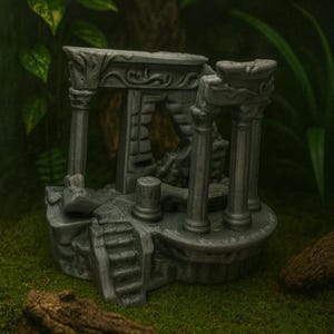 Op de afbeelding: Een grijze miniatuurreplica van een oude tempelruïne. De gedetailleerde structuur heeft kolommen, bogen en een kleine trap. Het model staat tegen een achtergrond van groen gebladerte en mos.