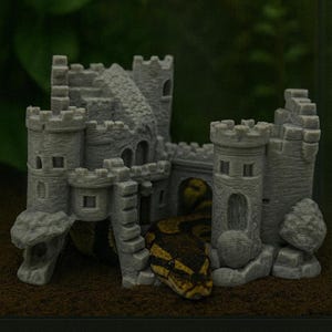 以下が含まれることがあります： 複数の塔と橋がある灰色の城の形をした水槽の装飾。茶色と黄色のヘビが部分的に見え、城の基部に巻き付いています。城はテクスチャ加工された表面を持っています。