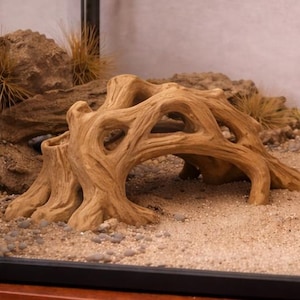 May include: A decorative, light brown, artificial driftwood structure with multiple archways, set in a terrarium with sand, small stones, and dried plants. The driftwood has a textured surface and is designed to mimic a natural habitat.
