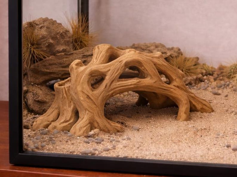 May include: A decorative, light brown, artificial driftwood structure with multiple archways, set in a terrarium with sand, small stones, and dried plants. The driftwood has a textured surface and is designed to mimic a natural habitat.