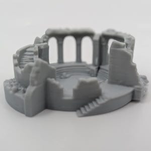 Può includere: Un modello in miniatura grigio di un'antica struttura in rovina. Il modello presenta un design circolare con colonne, scale e muri diroccati. L'estetica generale suggerisce un tema storico o fantasy, adatto per giochi da tavolo o esposizioni.
