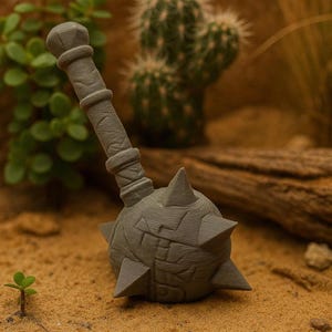 Puede incluir: Una maza gris impresa en 3D con un mango texturizado y una cabeza puntiaguda. La maza se encuentra sobre un fondo desértico con arena, plantas y un tronco. El diseño presenta tallados detallados y una estética medieval.
