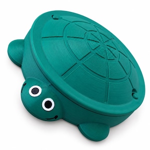 May include: A teal turtle-shaped container with a removable lid featuring a textured shell pattern. The turtle head has white circular eyes with black pupils and a small black smile. The body is a solid teal color with a smooth finish.