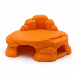 Könnte beinhalten: Ein leuchtend orangefarbenes, 3D-gedrucktes Versteck in Felsform mit einer flachen, erhöhten Plattform und zwei bogenförmigen Öffnungen an der Basis. Die Oberfläche hat eine texturierte, geschichtete Optik wie Stein.