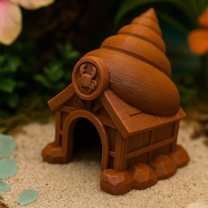May include: A brown, 3D-printed crab house with a spiral roof. The house features a circular emblem with a crab design and a rounded doorway. The structure is set on a sandy surface, suggesting a coastal or beach theme.