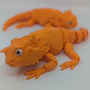 Peut inclure: Deux lézards jouets orange sont posés sur une surface blanche. Les lézards ont des membres articulés et des détails, notamment des yeux et une peau texturée. Un lézard est net, l'autre est légèrement flou en arrière-plan.