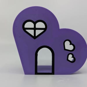 May include: A purple heart-shaped house with a black heart-shaped window and a black arched doorway. There are two smaller black hearts on the side of the house.