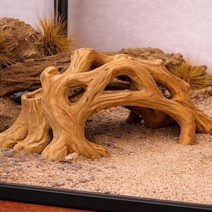 May include: A light brown, driftwood-style aquarium decoration. The artificial wood has a textured surface with multiple holes and arches, creating a naturalistic underwater habitat. The decoration sits on a bed of sand and small pebbles.