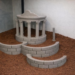 Puede incluir: Maqueta de hormigón gris de un templo clásico con columnas y techo parcialmente dañado. El templo se asienta sobre una estructura de ladrillo curvada y escalonada, rodeada de tierra marrón. Cerca hay un pequeño pilar rectangular.