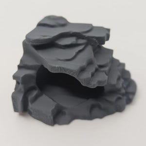 May include: A grey, layered rock formation with a cave-like opening. The rock has a rough, textured surface and appears to be made of a solid material. The design is reminiscent of a miniature landscape or a decorative element.