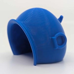 Puede incluir: Un objeto azul vibrante, impreso en 3D, con un diseño curvo único. Presenta una parte superior redondeada con un pequeño elemento que sobresale y un asa en forma de bucle en el lateral. El objeto tiene una superficie lisa y texturizada y una base abierta y arqueada.