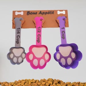 Puede incluir: Tres cucharas medidoras con forma de pata en gris, rosa y morado, colgadas de un estante de madera marrón. Las cucharas están etiquetadas con "1/4 Cup", "1/2 Cup" y "1 Cup". El estante tiene la frase "Bone Appétit" impresa.