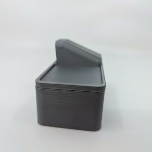 May include: A gray, rectangular box with a hinged lid and a small, angled top section. The box appears to be made of a solid material, possibly plastic or resin. The design is simple and functional, with clean lines and a matte finish.