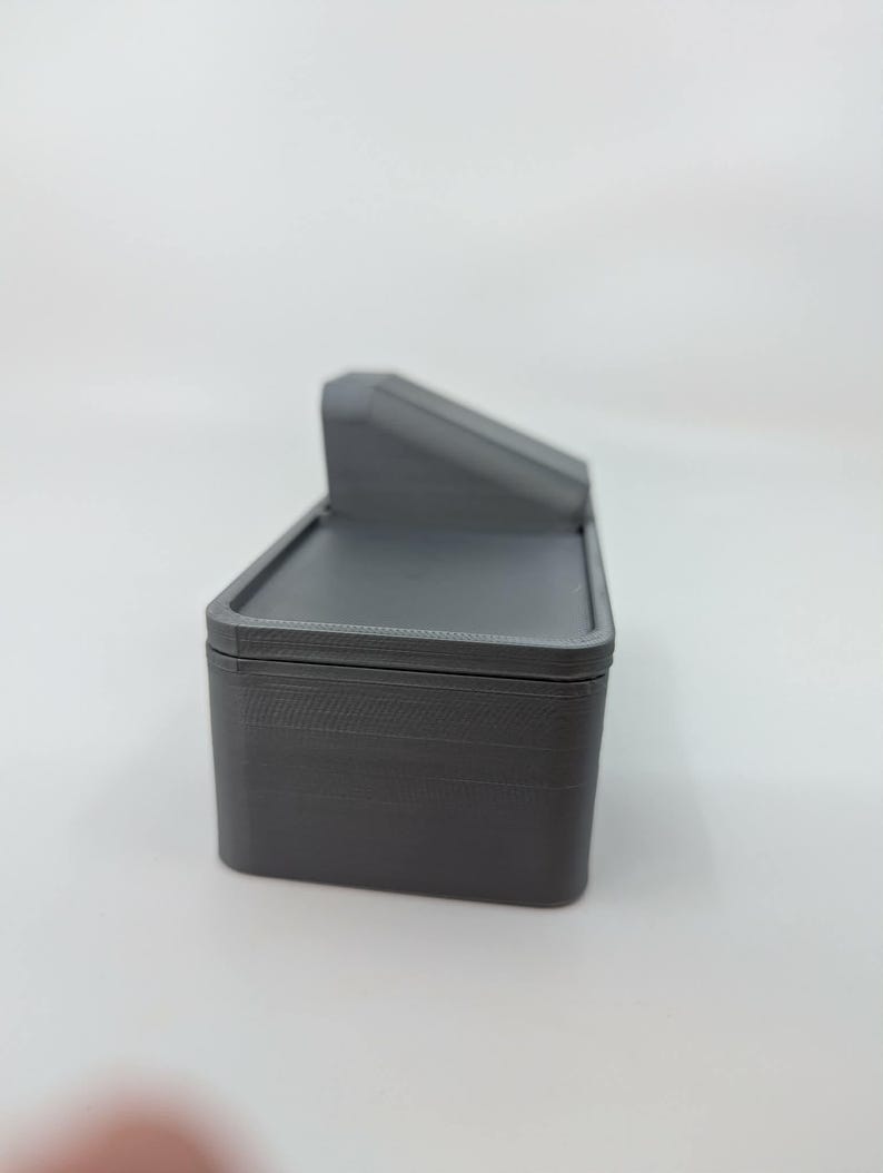 May include: A gray, rectangular box with a hinged lid and a small, angled top section. The box appears to be made of a solid material, possibly plastic or resin. The design is simple and functional, with clean lines and a matte finish.