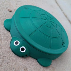 Puede incluir: Un juguete de tortuga de plástico verde con una concha texturizada y ojos saltones. La tortuga está diseñada para el juego de los niños.