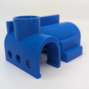 Op de afbeelding: Een helderblauwe 3D-geprinte speelgoedstructuur met een cilindrische toren, boogvormige openingen en een gebogen lichaam. Het speelgoed heeft drie ronde gaten aan de voorkant en een getrapt platform. Het ontwerp is modern en geometrisch.