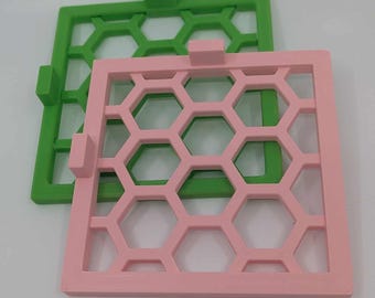 May include: Two square-shaped honeycomb-patterned stencils, one pink and one green. The stencils are made of a rigid plastic material.