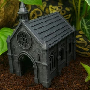Puede incluir: Una iglesia gris en miniatura con un diseño detallado, que incluye un campanario, una puerta arqueada y ventanas arqueadas. El modelo tiene una apariencia texturizada y está sobre un fondo natural con vegetación.