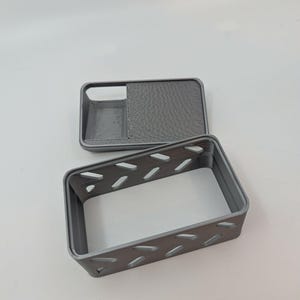 May include: A gray 3D-printed phone case with a matching rectangular box. The case has a textured back and a cutout for the camera. The box has a series of diagonal cutouts along its sides.