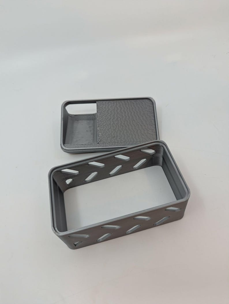 May include: A gray 3D-printed phone case with a matching rectangular box. The case has a textured back and a cutout for the camera. The box has a series of diagonal cutouts along its sides.