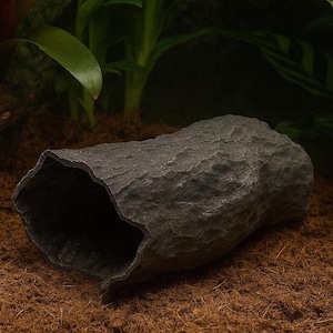 May include: A dark gray, rock-like reptile hideout with a hollow interior. The textured surface mimics natural stone, providing a secure and visually appealing habitat accessory. The hideout is designed to blend seamlessly with a terrarium environment.