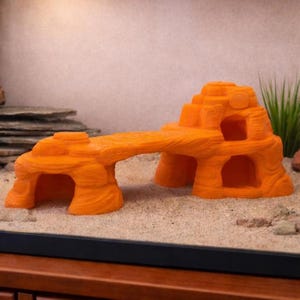 May include: Orange aquarium ornament designed as a rock formation with a bridge and openings. The textured surface and colour create a natural appearance for aquatic environments. The ornament is placed on a sandy substrate.