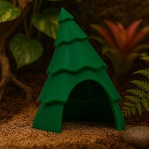 Può includere: Un rifugio verde a forma di albero per piccoli animali domestici. Il rifugio ha una forma triangolare con un'apertura arrotondata e lati strutturati a strati. È posto su un letto di sabbia, con fogliame sullo sfondo.