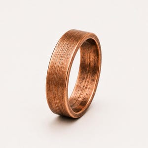 Puede incluir: Un anillo de cobre con un acabado cepillado y texturizado en la superficie exterior. La banda presenta un tono marrón rojizo metálico cálido con finas estrías verticales a lo largo de la circunferencia, creando un aspecto desgastado.