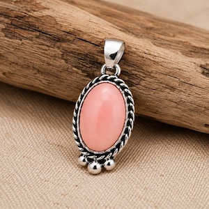 May include: An oval pendant featuring a smooth, light pink cabochon stone set in a polished silver frame. The setting is decorated with a twisted rope border and three small silver spheres at the base, topped with a silver bail for hanging.