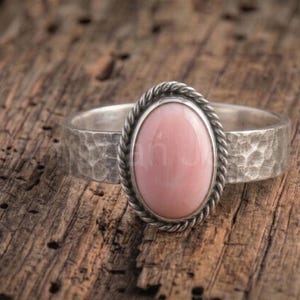 Könnte beinhalten: Ein Silberring mit einem glatten, ovalen rosa Stein, der von einer gedrehten Kordelbordüre eingefasst ist. Die Ringschiene weist eine texturierte, gehämmerte Oberfläche auf, die einen deutlichen Kontrast zur polierten Fläche des Steins bildet.