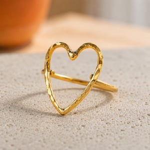 May include: A gold-toned ring featuring an open heart-shaped design. The metal surface of the heart outline has a distinct hammered texture, creating a faceted appearance that reflects light across the thin, circular band of the ring.