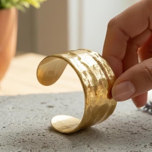May include: A gold-colored cuff bracelet with a textured, hammered surface. The interior of the open-ended band features a clear stamp reading 925, indicating the metal composition. The bracelet has a wide, curved design with polished edges.