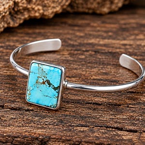 May include: A silver cuff bracelet featuring a rectangular turquoise stone with dark brown matrix veining. The polished metal band is open at the back, showcasing a smooth, reflective finish that contrasts with the vibrant blue of the stone.