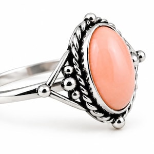 Puede incluir: Un anillo de plata con una piedra ovalada lisa de color melocotón engastada en un marco de metal trenzado. El engaste está decorado con pequeñas esferas de plata en los laterales, unidas a una banda de plata pulida y delgada.