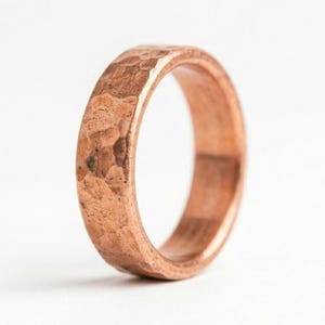 Op de afbeelding: Een koperen ring met een getextureerde, gehamerde afwerking aan de buitenzijde. De band heeft een constante breedte en een warme, metalliek roodbruine kleur. De binnenzijde van de ring is glad, terwijl de buitenzijde onregelmatige deukjes vertoont.