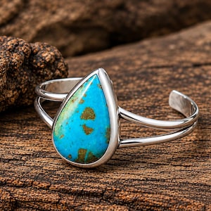 May include: A silver cuff bracelet featuring a large, teardrop-shaped turquoise stone with brown matrix inclusions. The band is constructed from two smooth, polished silver wires that meet at the setting of the vibrant blue-green gemstone.