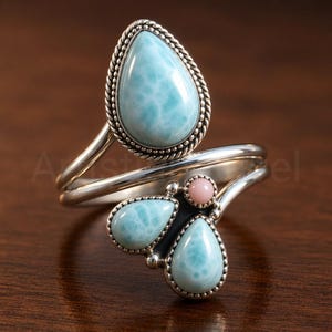 Op de afbeelding: Een open ring van sterling zilver met een grote, peervormige lichtblauwe Larimar-steen met een gedraaide rand, en twee kleinere peervormige Larimar-stenen gecombineerd met een enkele kleine ronde roze steen aan het andere uiteinde van de ring.