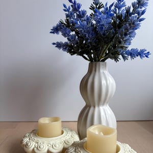 May include: Two white ceramic candle holders with intricate designs, each holding a lit ivory candle. A white vase with a textured surface holds a bouquet of blue lavender.