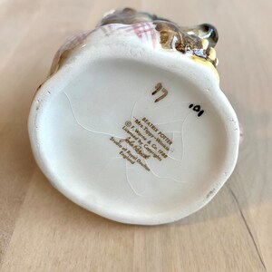 May include: White ceramic figurine of Beatrix Potter's Mrs. Tiggy-Winkle, a hedgehog, wearing a red and white checked dress. The figurine is sitting on a base with the text "Beatrix Potter", "Mrs. Tiggy-Winkle", "F. Warne & Co. 1988", "Licensed by Copyrights", "John Berk", "Studio of Royal Doulton", and "England".