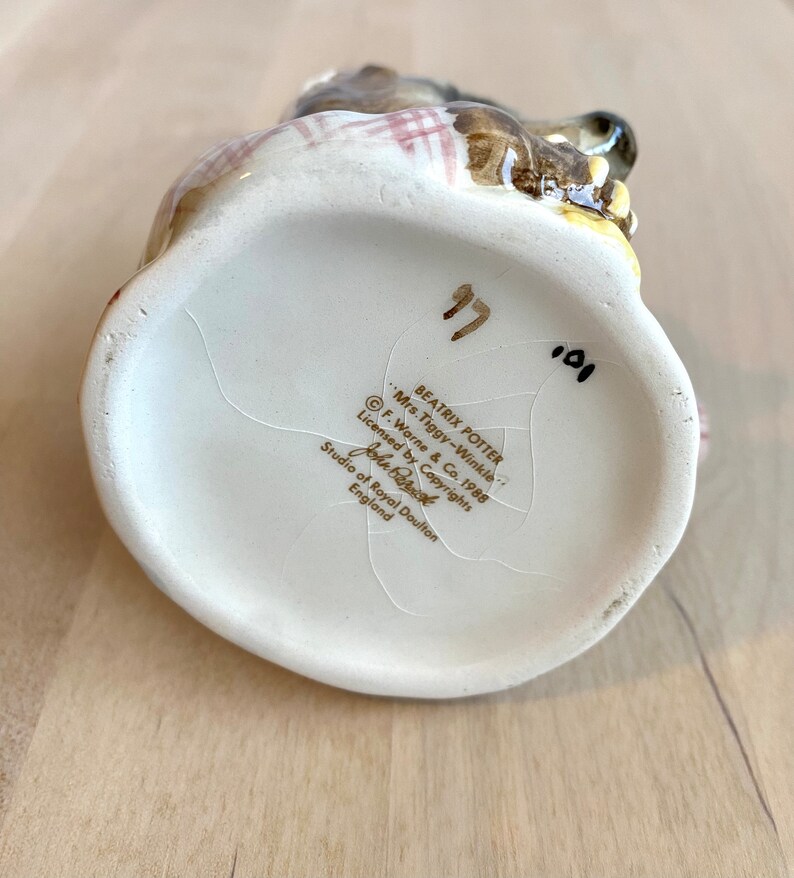 May include: White ceramic figurine of Beatrix Potter's Mrs. Tiggy-Winkle, a hedgehog, wearing a red and white checked dress. The figurine is sitting on a base with the text "Beatrix Potter", "Mrs. Tiggy-Winkle", "F. Warne & Co. 1988", "Licensed by Copyrights", "John Berk", "Studio of Royal Doulton", and "England".