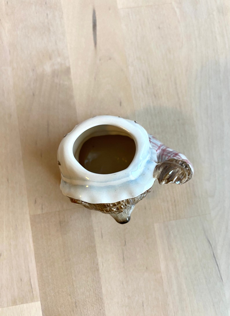 May include: A ceramic mug shaped like a brown bear wearing a white and red checked scarf. The mug is empty and the inside is visible.