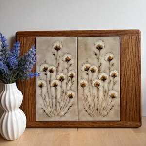 May include: A decorative wooden framed tile art piece featuring a floral design. The artwork has two beige tiles with a repeating pattern of white and brown flowers. A white vase with blue flowers is to the left.